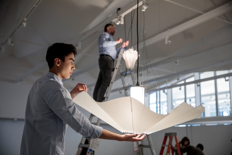 Person in foreground supports a paper chandelier while another person in the background on a ladder hangs a conical lamp from the ceiling of a gallery space.