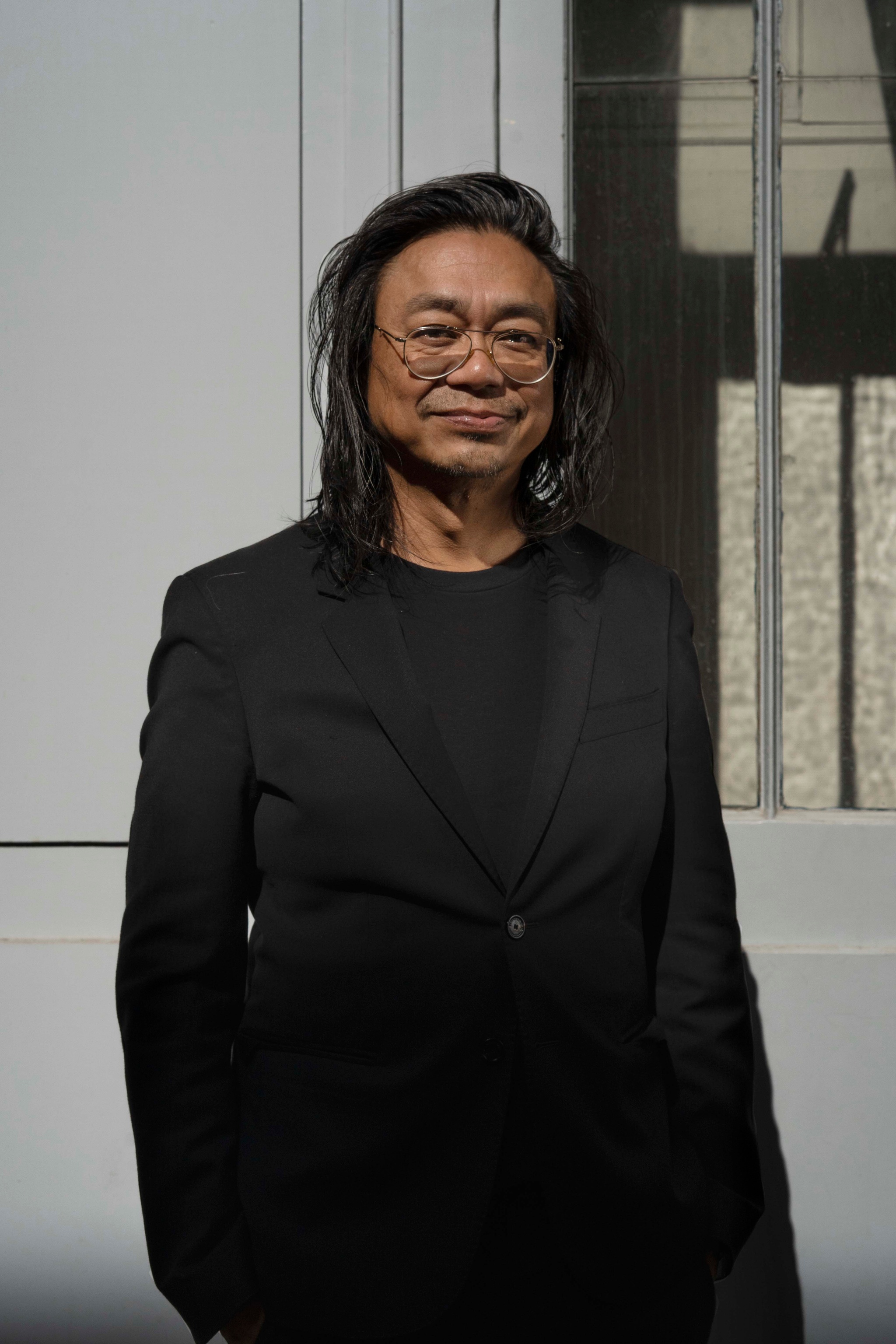 A Thai man with shoulder length hair and wearing glasses smiles wryly. He stands with arms at his side against a concrete wall.