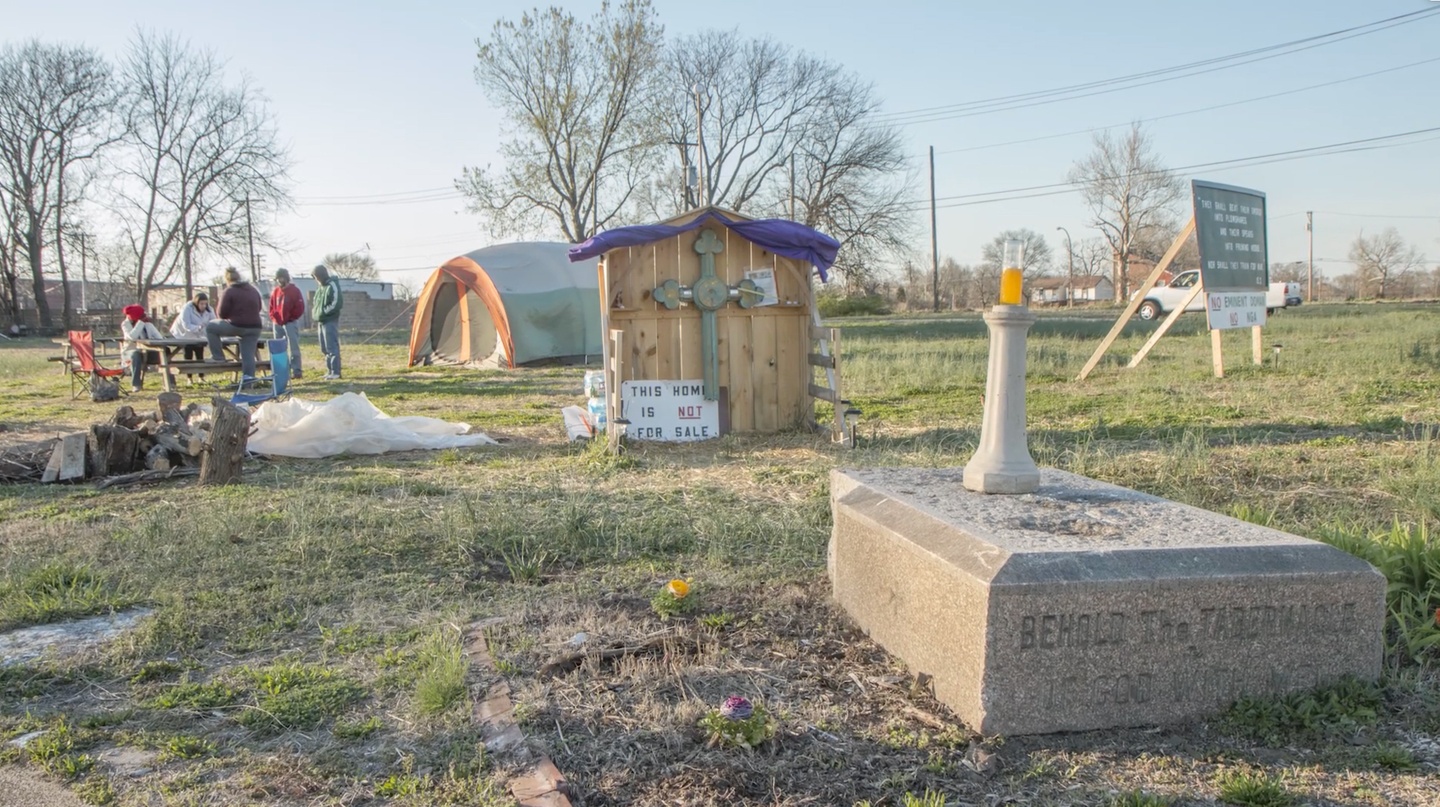 Film still showing people on a grassy lot, seated at a picnic table, with a camping tent to their right. In the foreground is a memorial stone commemorating a church that had previously exited on the site, as well as a wooden memorial with a large cross and a sign that reads "This home is not for sale."