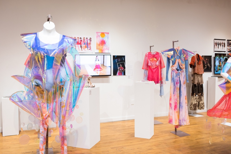 View of a fashion design collection. The garment in the foreground is made of iridescent plastics arranged in upturned ruffs with a bralette and colorful leggings underneath.
