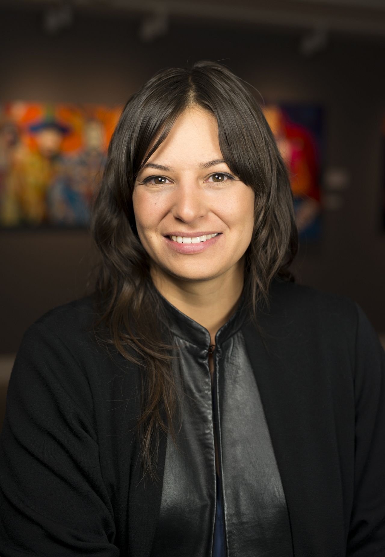 A portrait of Candice Hopkinss, a citizen of Carcross/Tagish First Nation. She has dark brown hair parted at the center of her forehead that falls below her shoulders. She wears a black jacket with a wide lapel that has the texture of leather. She smiles with a blurry artwork behind her in the background. 