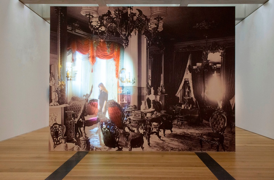 A large-scale screen, hung from the ceiling, featuring an ornately decorated room that's mostly black-and-white, except for a circular section to the left that is full-color and somewhat blown-out through the window area, showing a person standing in front of the windows.