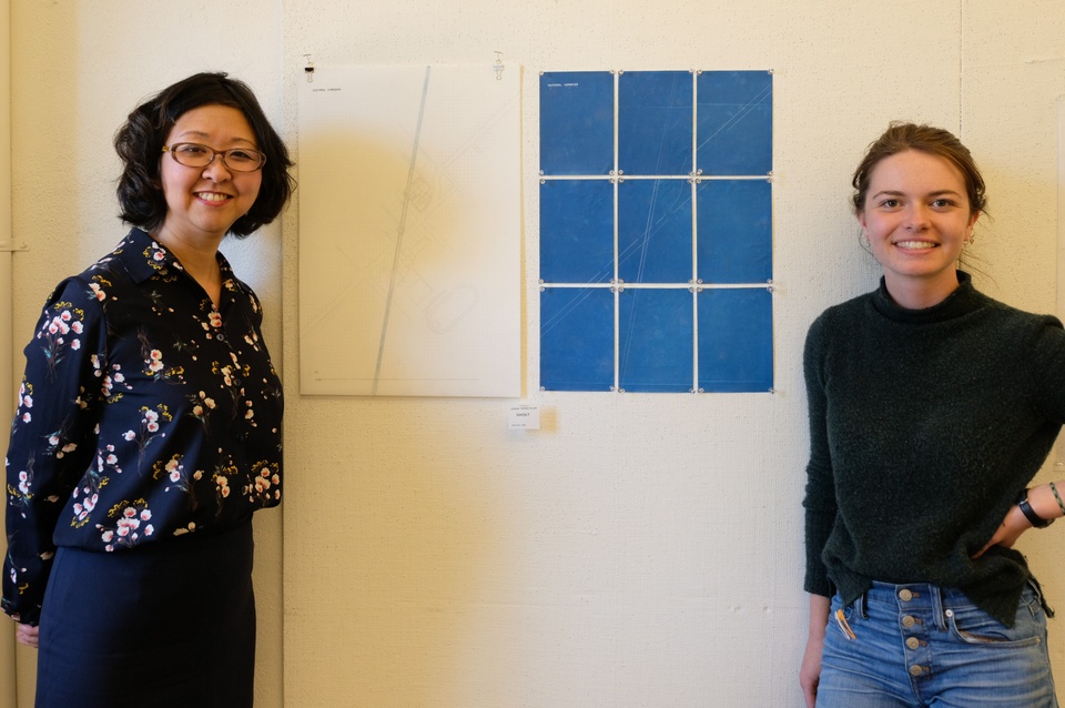 A visiting faculty and a student stand smiling at the camera, with the student's pinned up architectural drawing on the wall between them.