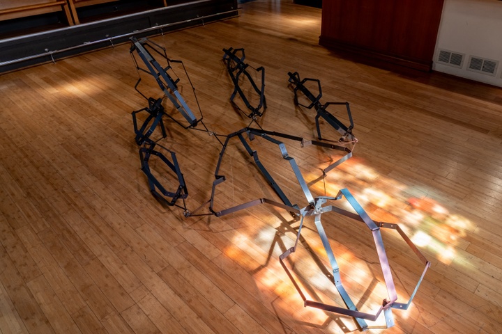 Large metal sculpture made of crimped black metal strips bent in symmetrical shapes and connected to each other by joints lying on the floor.