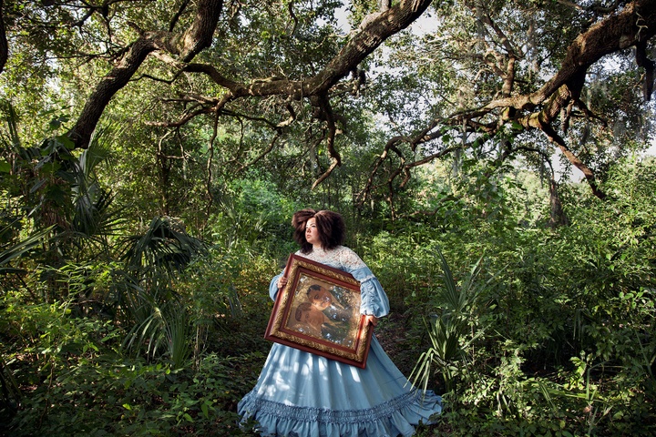 Full-color inkjet print featuring in an elaborate, light-blue, Victorian-era dress, walking through a tree-filled forest carrying a painting.