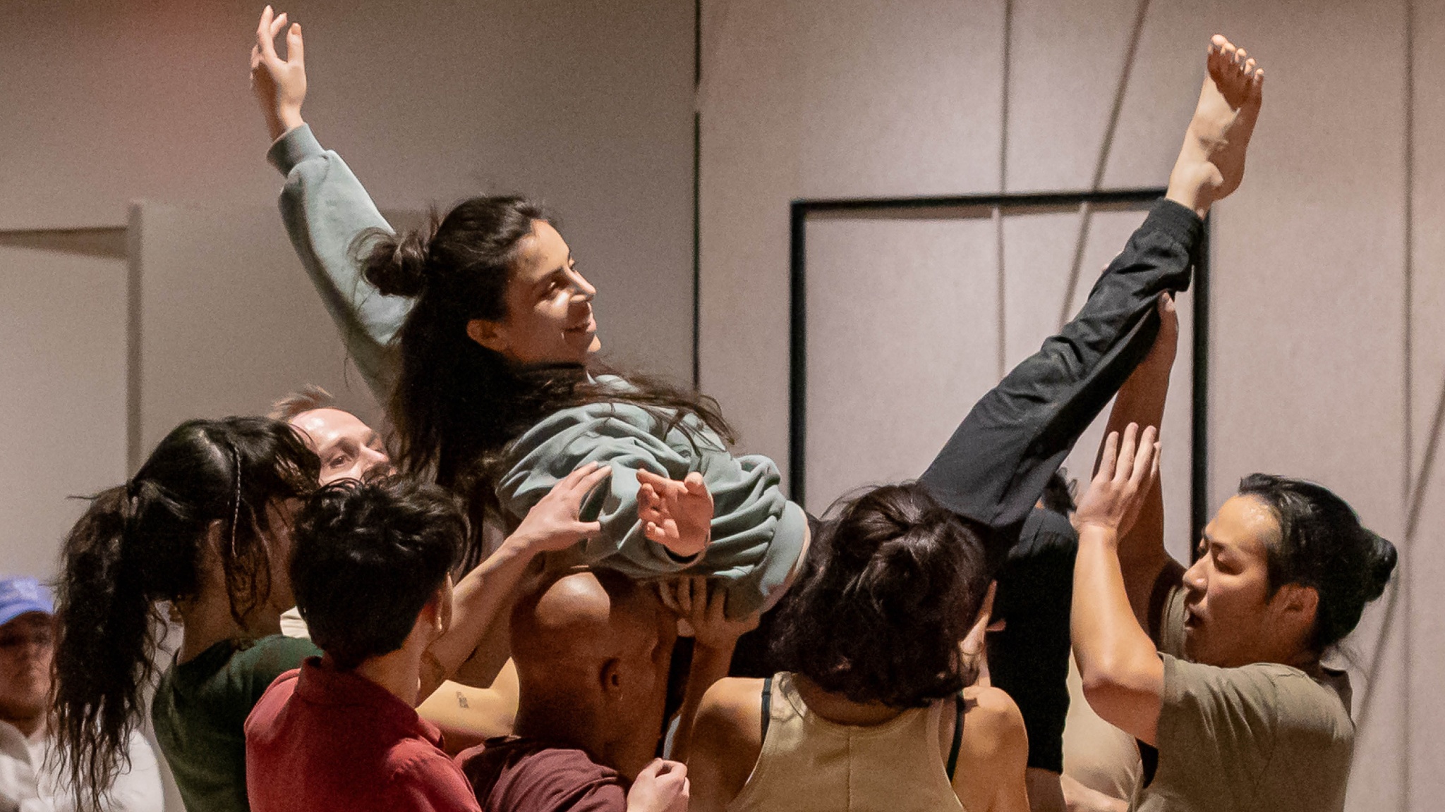 An ensemble of dancers lift another performer overhead. The dancer being held aloft wears a green sweatshirt and gray sweatpants with her arms outstretched exuberantly, extending one leg with a bare foot pointed toward the ceiling. She is smiling while the other performers support her.