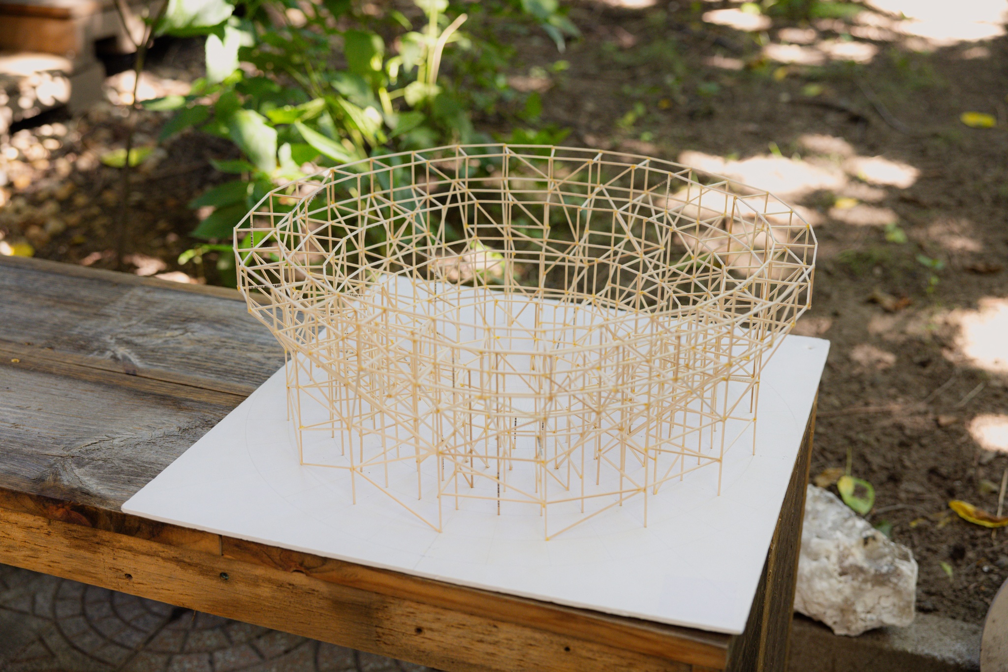 A scale model made of thin toothpick like sticks forms the circular structure of a cockfighting ring. It sits on a wooden table in a shady backyard. 