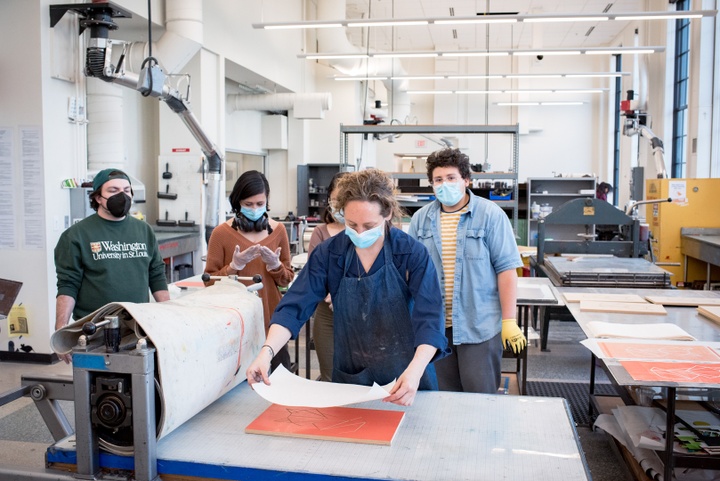 Group of people in a printmaking studio. One person carefully lays a sheet of paper down on an orange-inked relief print block on a press table.