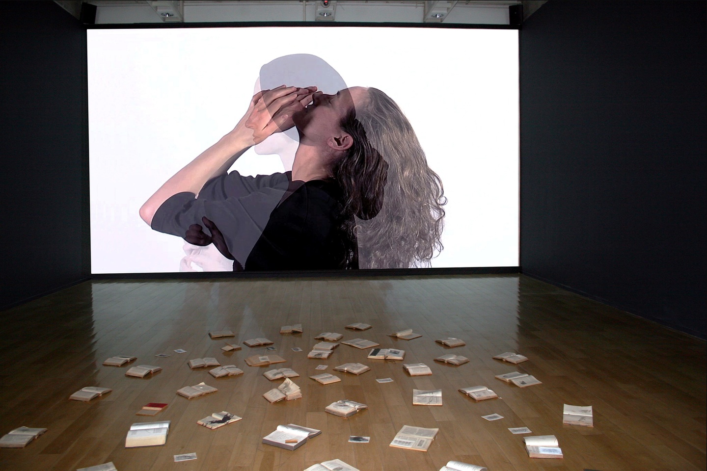Installation view of a gallery: in the center, on the wall is a projection of a video; in this video are two figures in profile, overlapping; one person arches their head back, their hands holding their face; the overlaid image is of a person standing upright. The background of the video is white. Before this video in the foreground are scattered books, all laid out in open spreads. The lighting is dim in the background, illuminating the books closest to us.