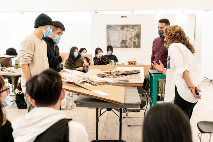 Two students tilt a cardboard city plan model up so that two instructors can see and comment on it.