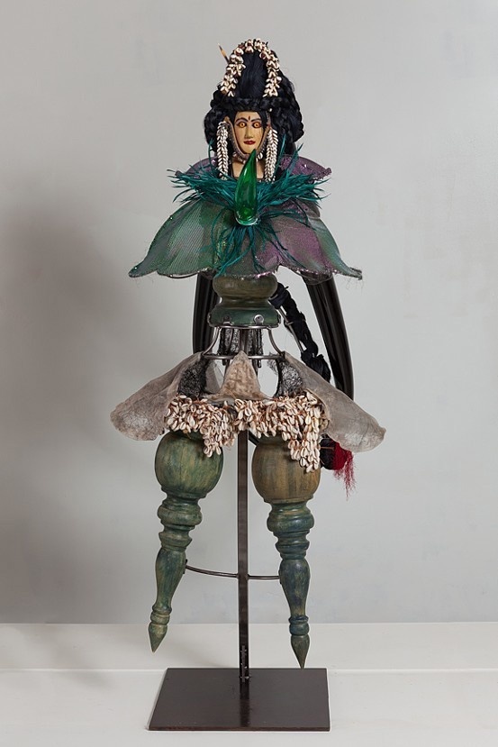 A sculpture of an abstract carved figure with a flared dress and cape made of found material. The figure wears an elaborate, black wig. 