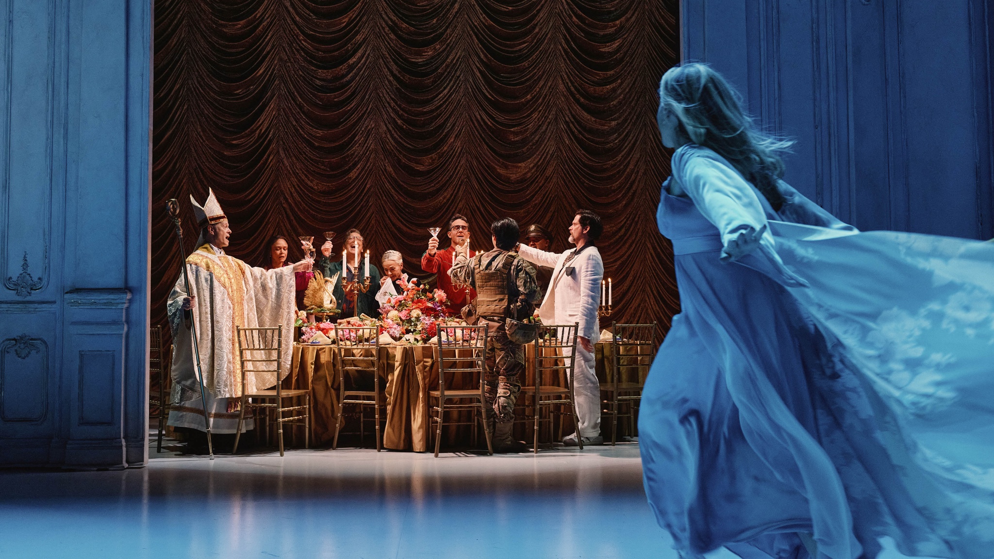 The cast of a play dressed as members of a dinner party lift glasses in a toast as they stand around a long table. At one end stands a guest dressed in white Catholic clerical robes and at the other end a man in a white suit stretches his arm out. In the foreground lit in liquidy blue light a white woman in a nightgown runs toward them, her gown and blond hair flowing behind her. 