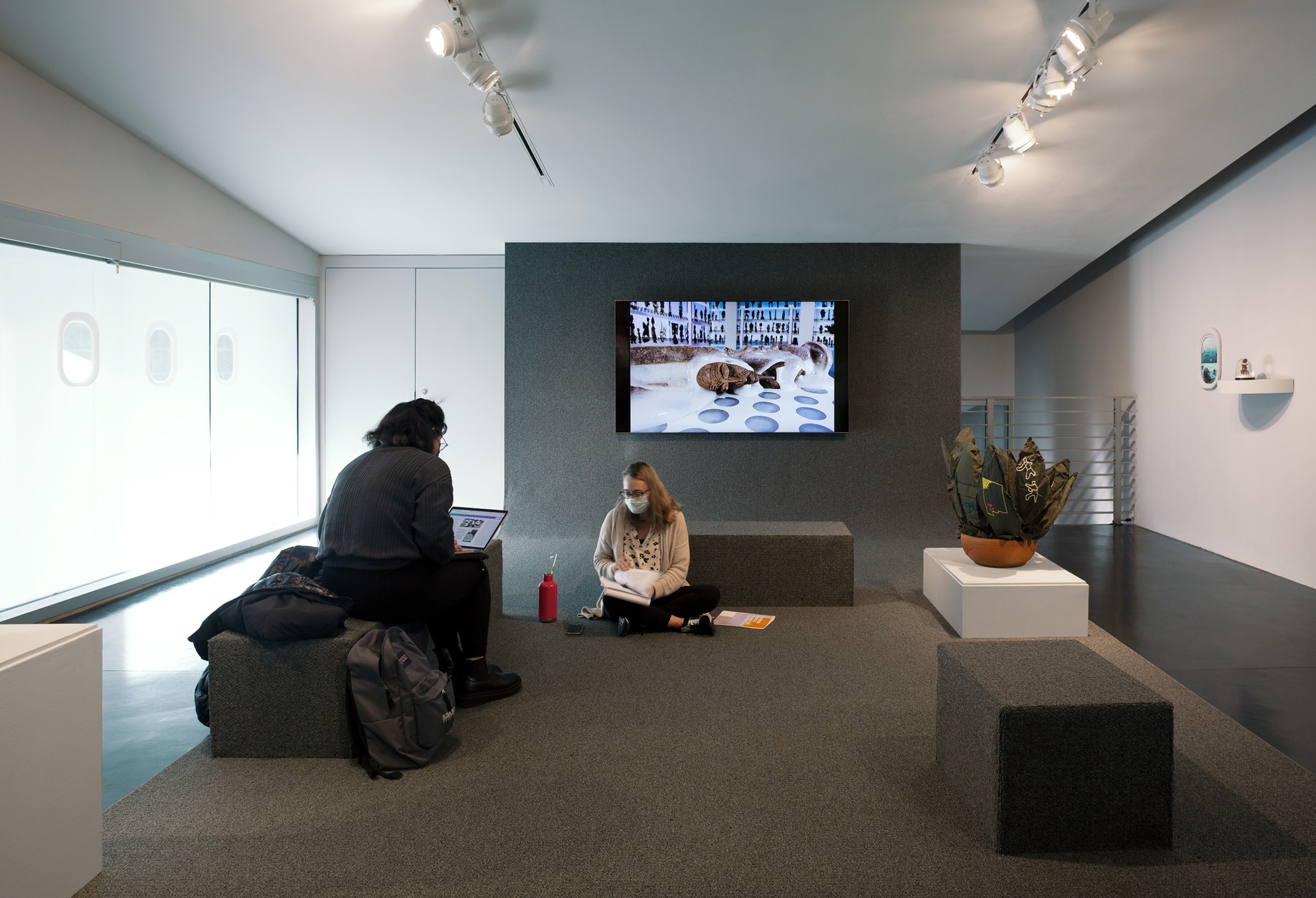 Two people sitting in a gallery with a soft sculpture of a cactus to their right and a television monitor of an animated image on the wall behind them.
