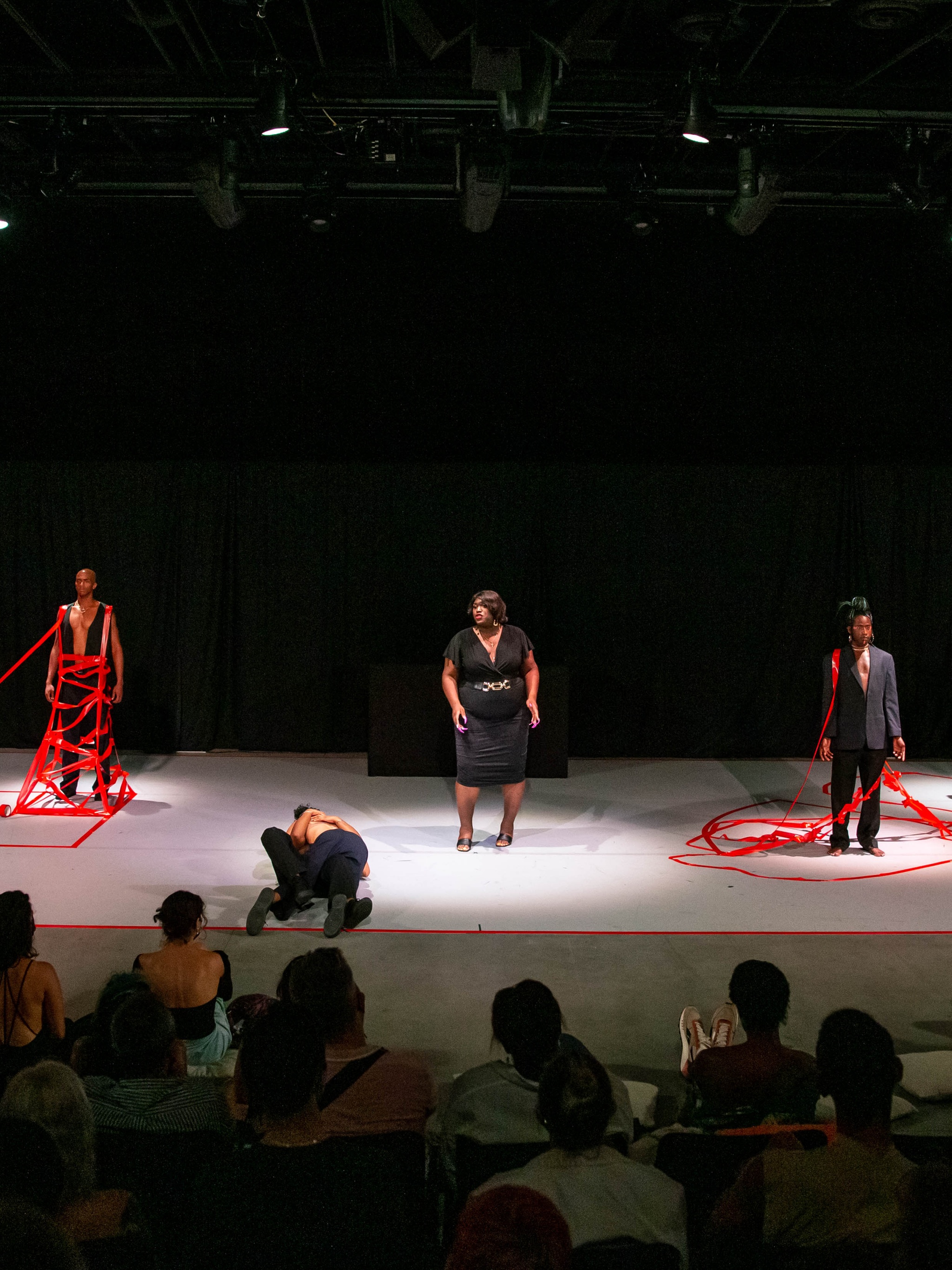 Three Black trans performers stand on a stage wearing black suits and dresses in front of an audience. On the floor of the stage, two performers embrace as they roll together across the floor. Two of the standing performers have red tape reaching up from the ground to hold them in place.