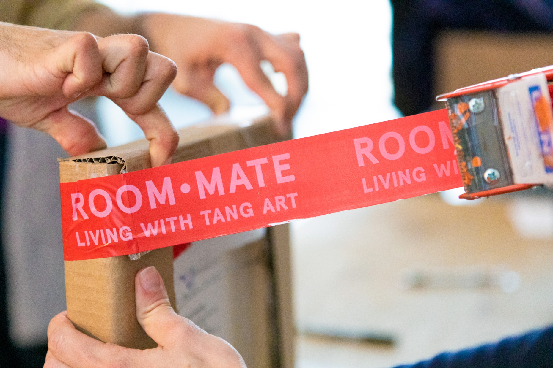 Red tape that reads “ROOMMATE LIVING WITH TANG ART” is being taped onto a box where two hands hold the top of the box and one hand on the side.