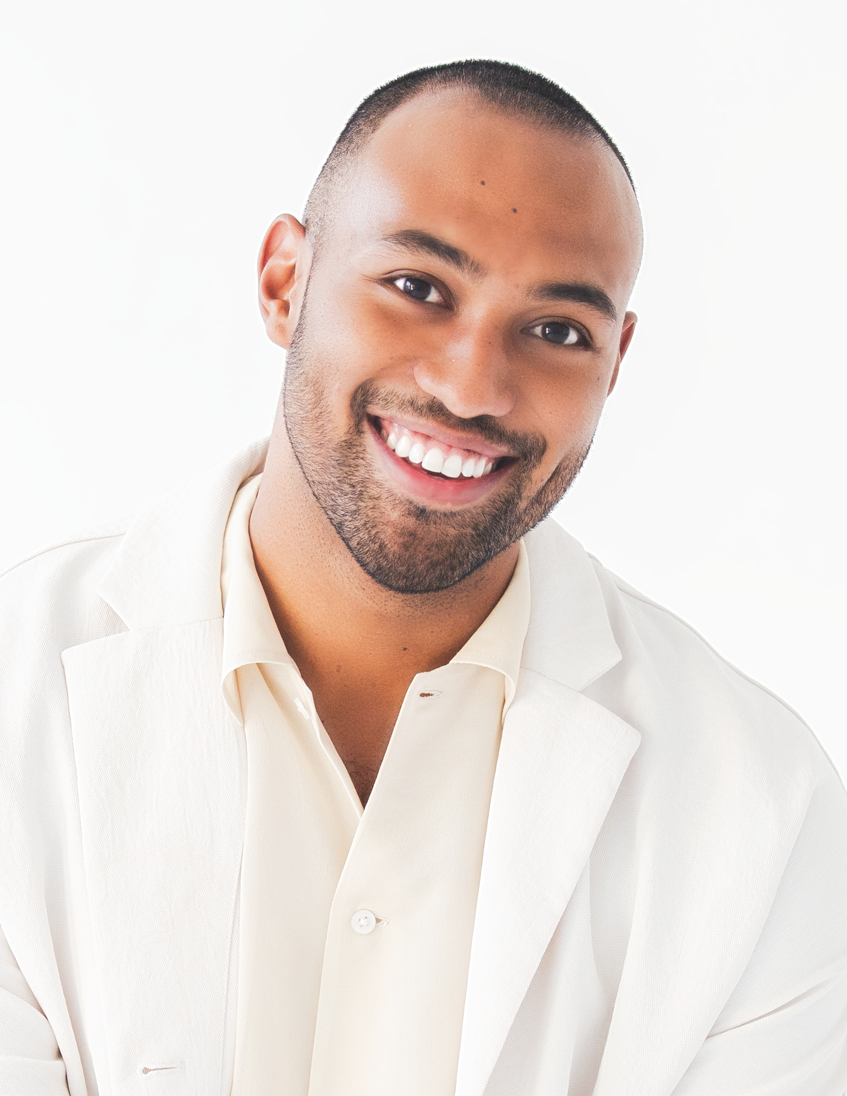 A portrait of Adam Hyndman who poses in a white shirt against a white background. He has light brown skin, a light beard, and smiles broadly at us.