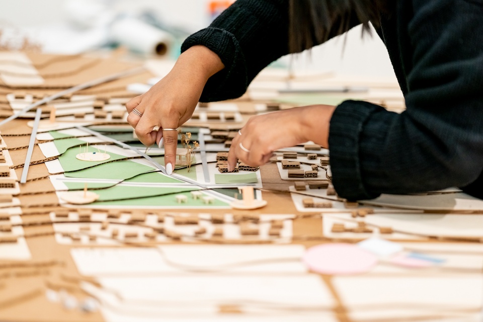 Two disembodied hands are adding green paper to a city plan model's park area.