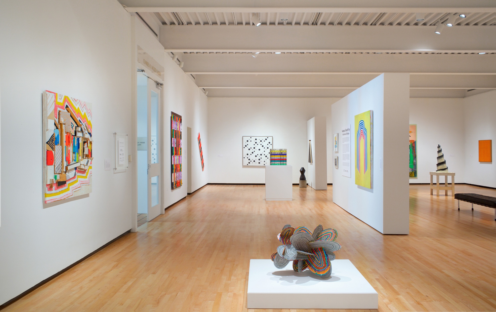 Bright, abstract artwork hangs in a gallery with white walls and wood floors with sculptures placed centrally throughout.