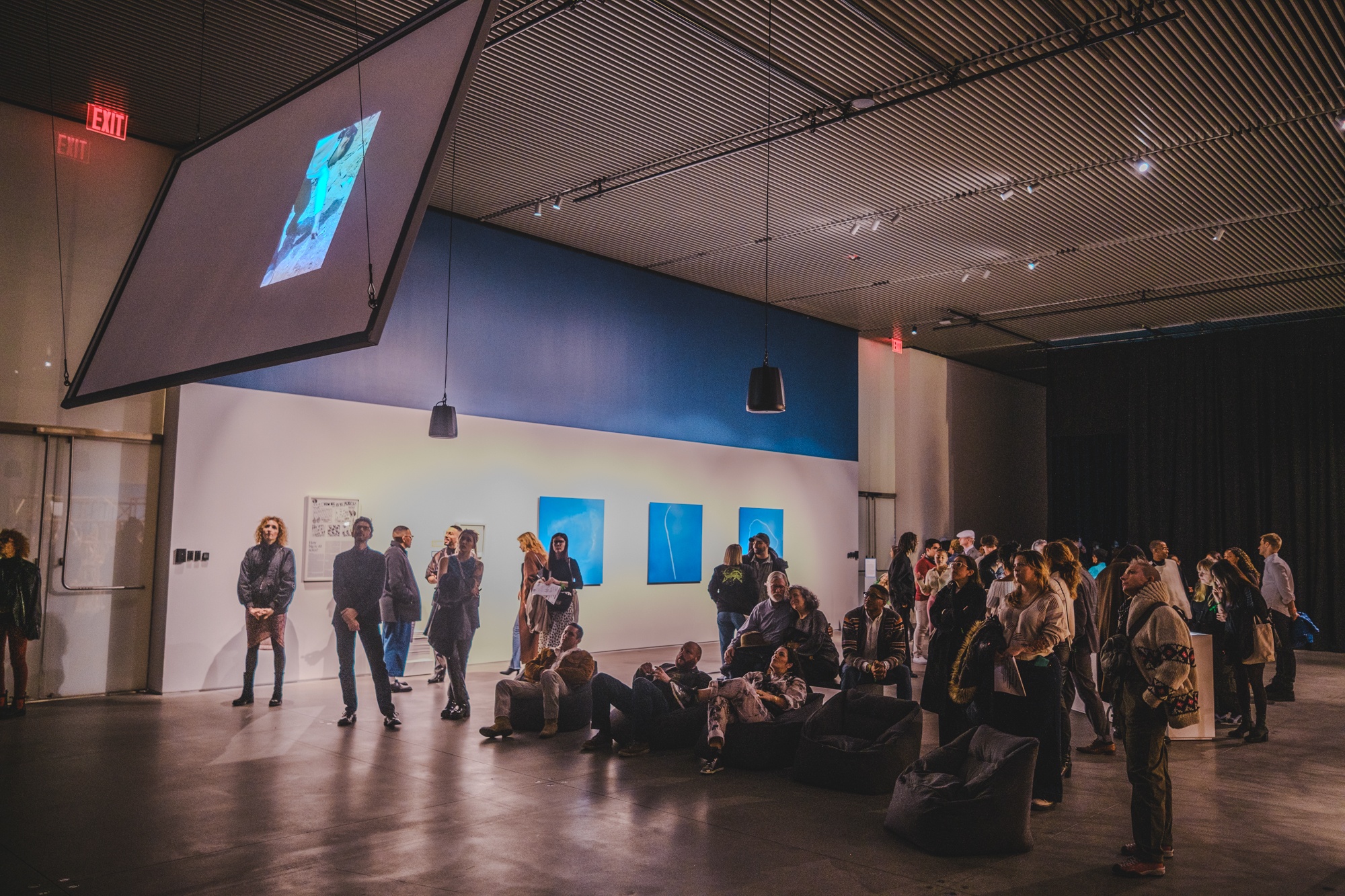 An audience gathers in a gallery standing and sitting on bean bag chairs beneath a film screen that hangs from the ceiling on a diagonal. They look up at the screen to watch a film. Behind them, three photographic prints of clouds created by a skywriting plane hang on the wall.