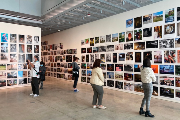 In an airy, contemporary gallery space, individuals view walls lined floor to ceiling with unframed and unmounted photographs.