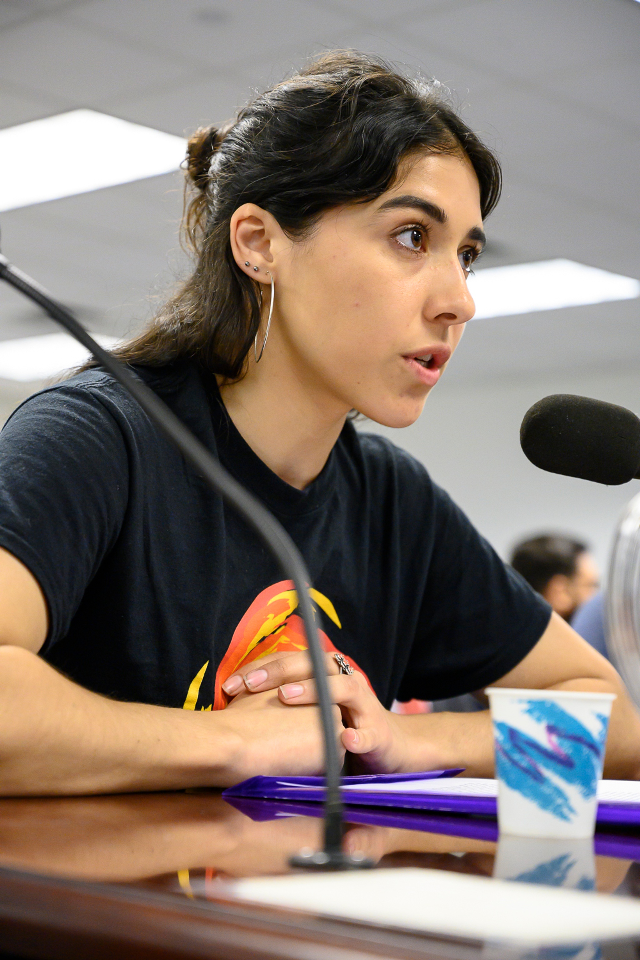 A photo of Leslie Velasquez speaking into a microphone at a table with her elbows on the table and hands crossed in front of her