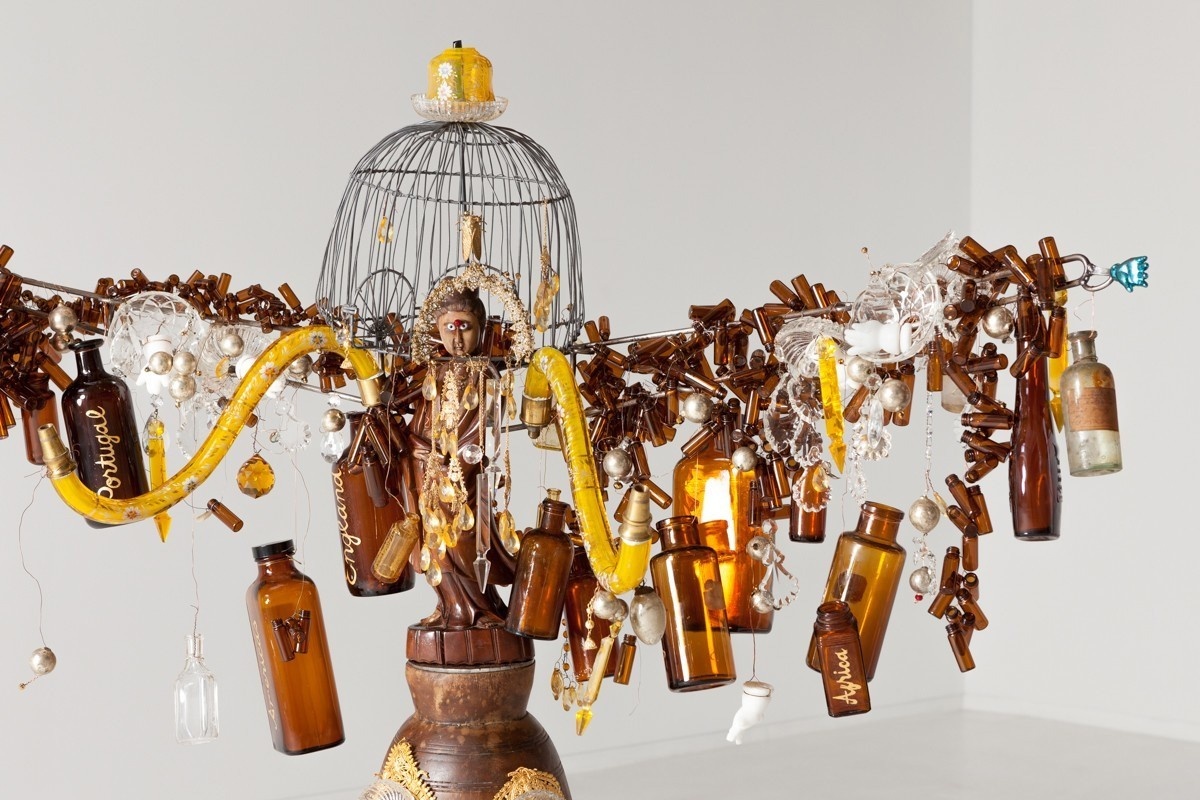 A carved, brown figure stands under a metal domed cage in the center of numerous outstretched arms with brown glass bottles.