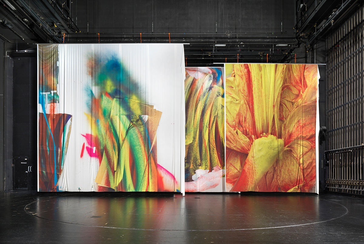 Three printed fabric screens hanging in a darkened stage-like space, featuring fragments of abstract paintings
