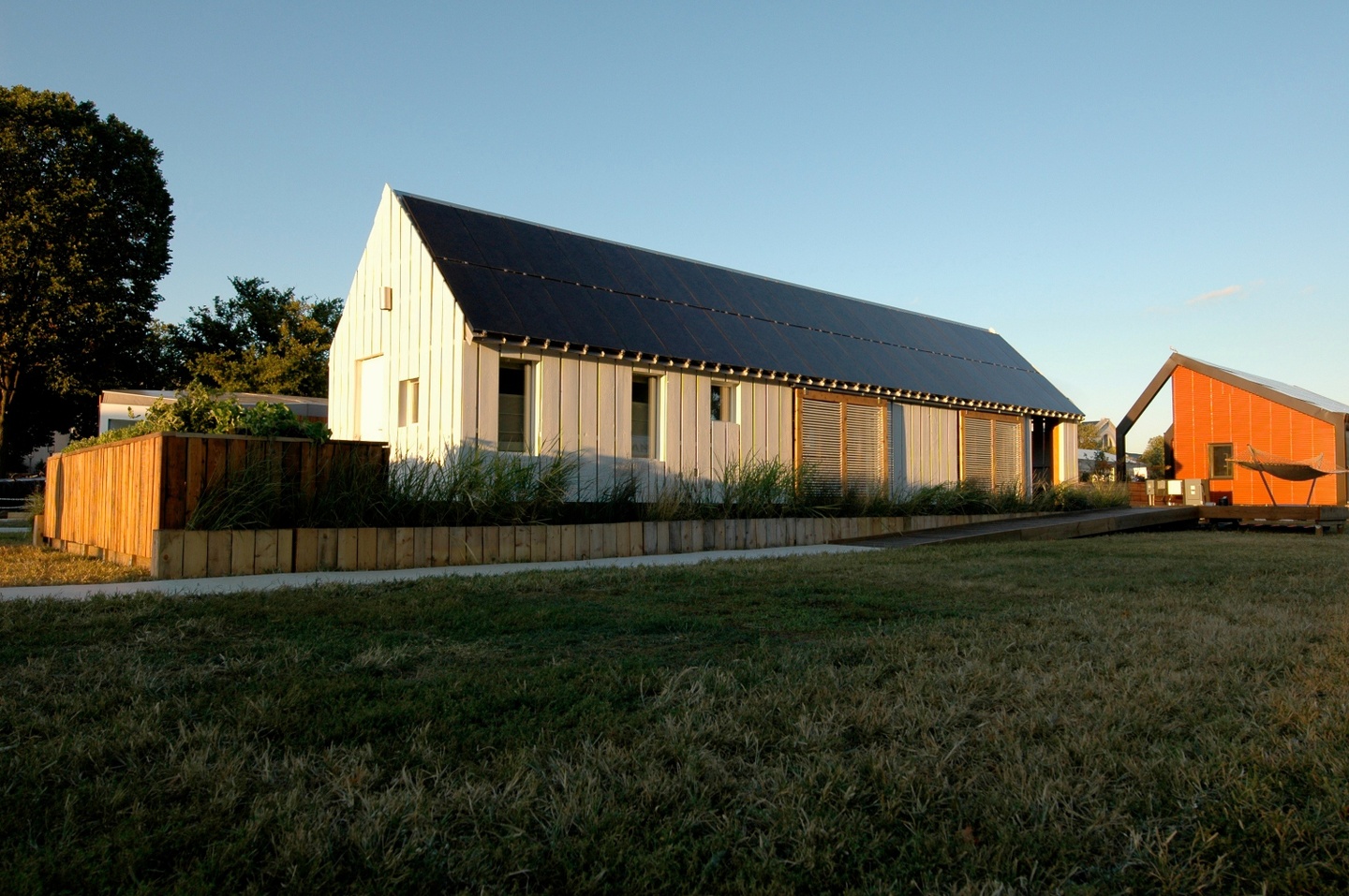 Single-story gabled house with white vertical siding panels and a solar panel roof, surrounded by wooden tiered garden boxes.