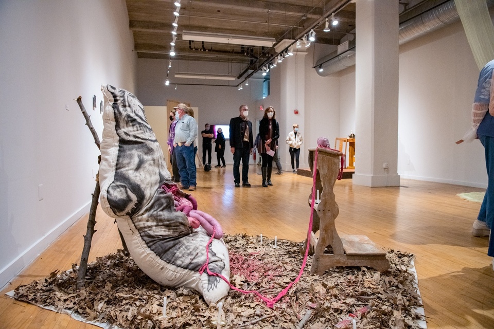 Small gallery space. In the foreground is a 4x4' square carpet of dead leaves. Propped against two sticks is a large pillow in the shape of a bear with a bear imprint on the front. Its stomach is sliced open and fabric entrails are spilling out.