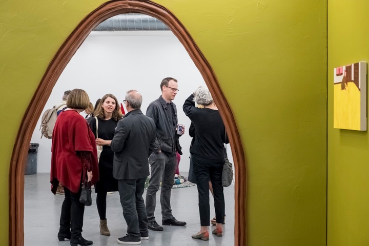 3D sculpture featuring two chartreuse walls; the right wall has a small artwork hung on it, while the back wall has an archway cut out, with people in view through it.