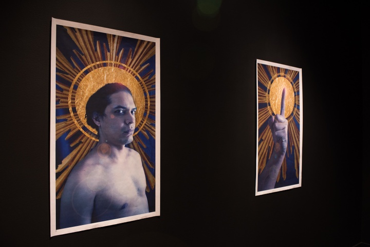 Two prints on a black wall. The left depicts a shirtless person, torso up, looking at the viewer, with a saintly halo behind their head. The right print is a clenched fist with the pointer finger upwards, also against a saintly halo.