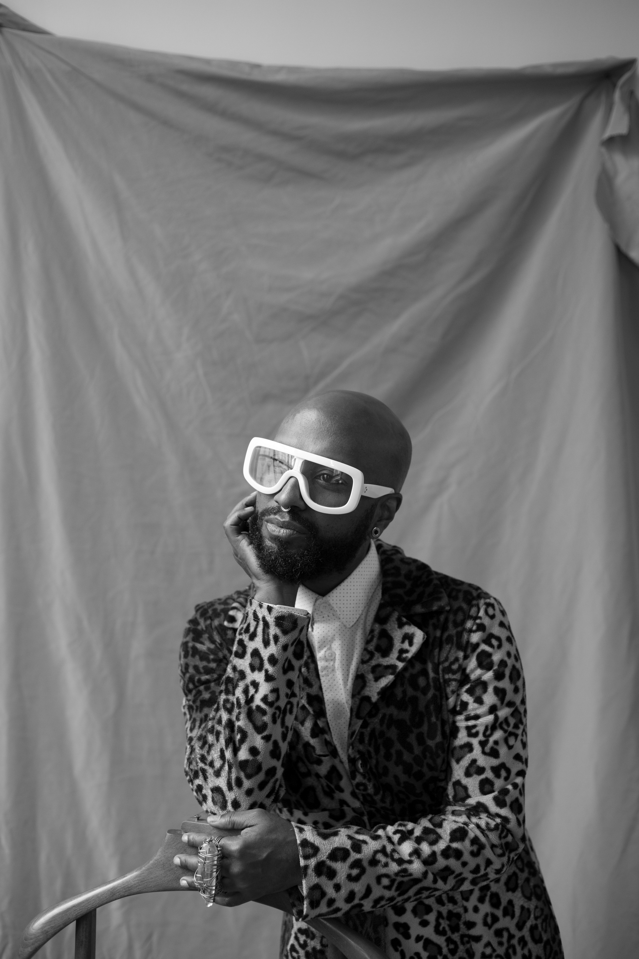 A portrait of Monstah Black, a Black artist who has a shaved head and wears oversized square frame white glasses. Monstah poses against a fabric backdrop with one hand supporting his chin. He wears a leopard print blazer. Photo by Lia Clay. 
