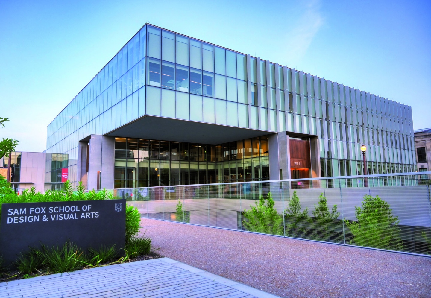 A glass and metal academic building reflects the blue, early-dawn light. In the foreground, a dark gray granite sign reads Sam Fox School of Design & Visual Arts. 