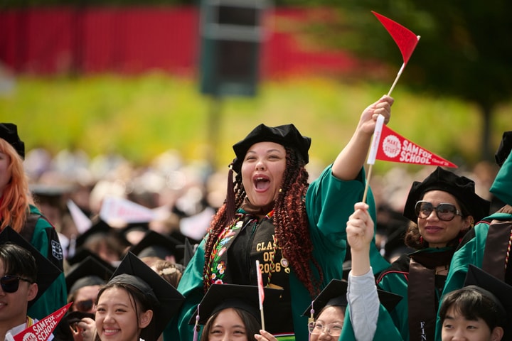 A student wearing regalia and waving a pennant stands and cheers