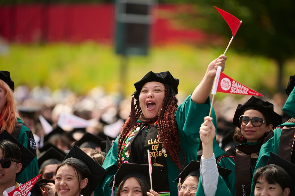 A student wearing regalia and waving a pennant stands and cheers