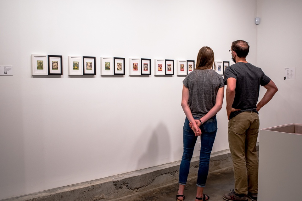 Two people in masks look at a row of framed playing card designs.