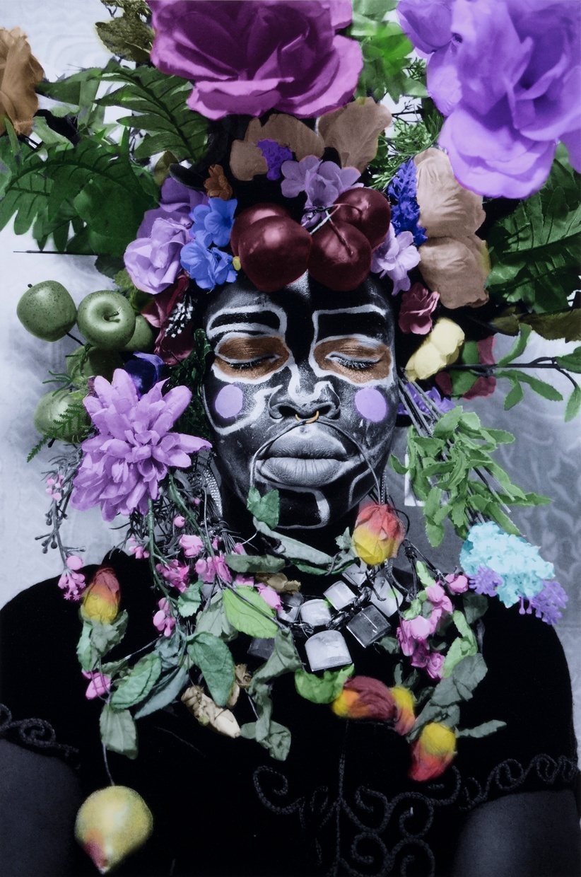A figure with closed eyes faces forward, their face painted in black and white and colorful flowers and fruit surround their head.