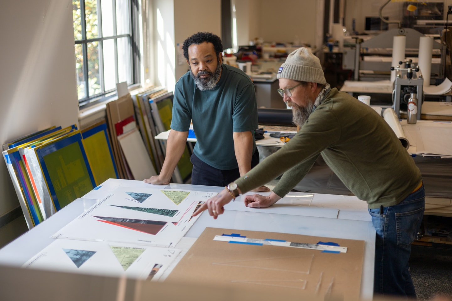 two men standing at a table in a printmaking studio. The man on the left has his hands resting on the table while listening to the man on the right as he points to the print