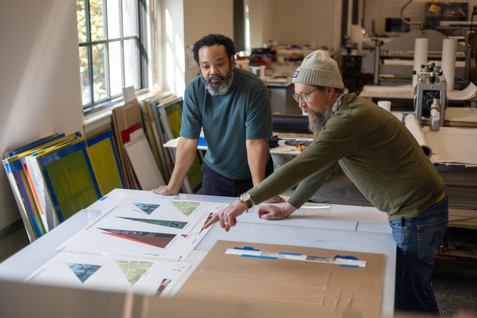 two men standing at a table in a printmaking studio. The man on the left has his hands resting on the table while listening to the man on the right as he points to the print
