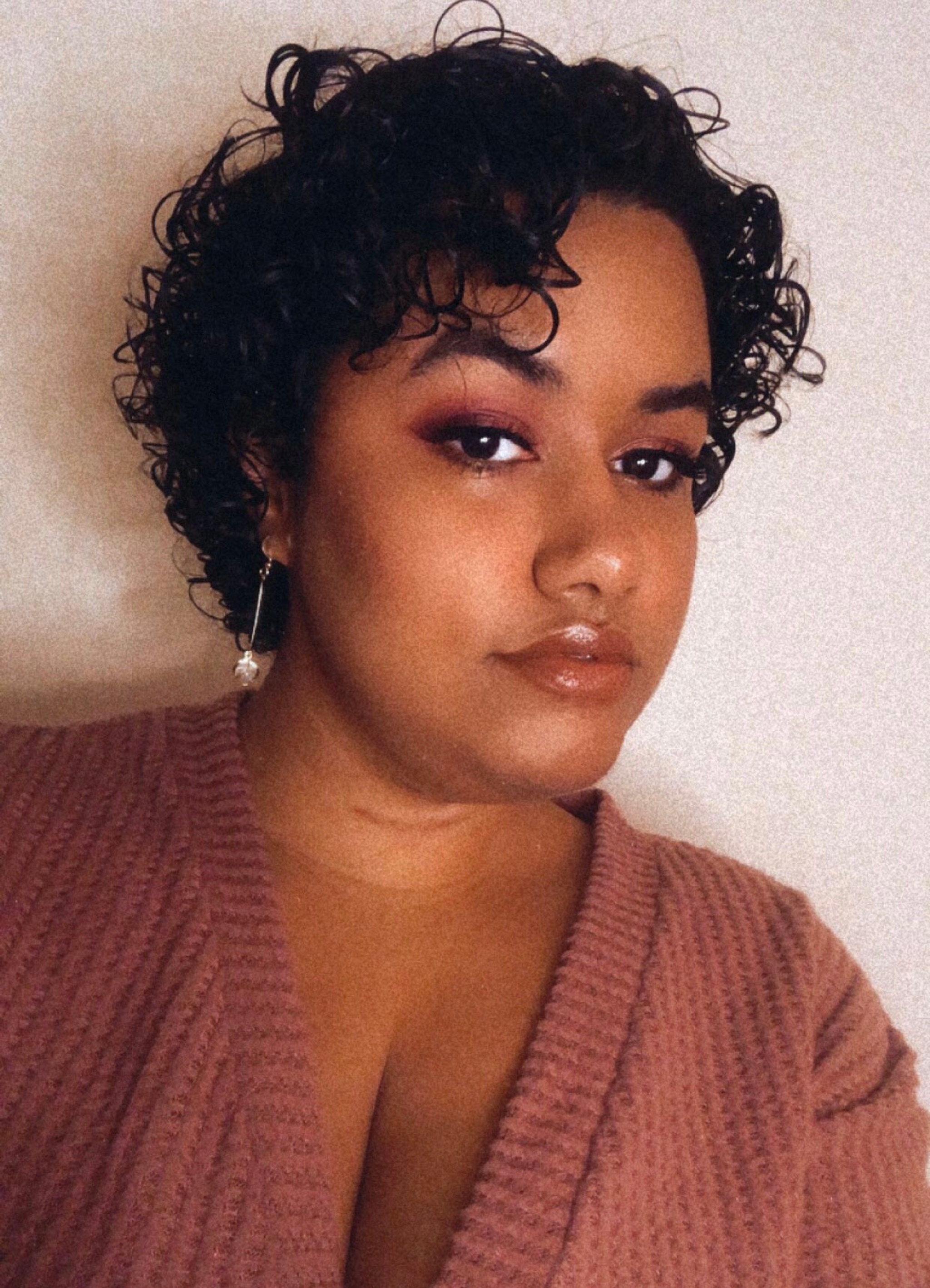 A portrait of singer Genesis Adelia Codallo, a Dominican person who has short curly hair that falls in rings over her forehead and wears a light pink sweater.