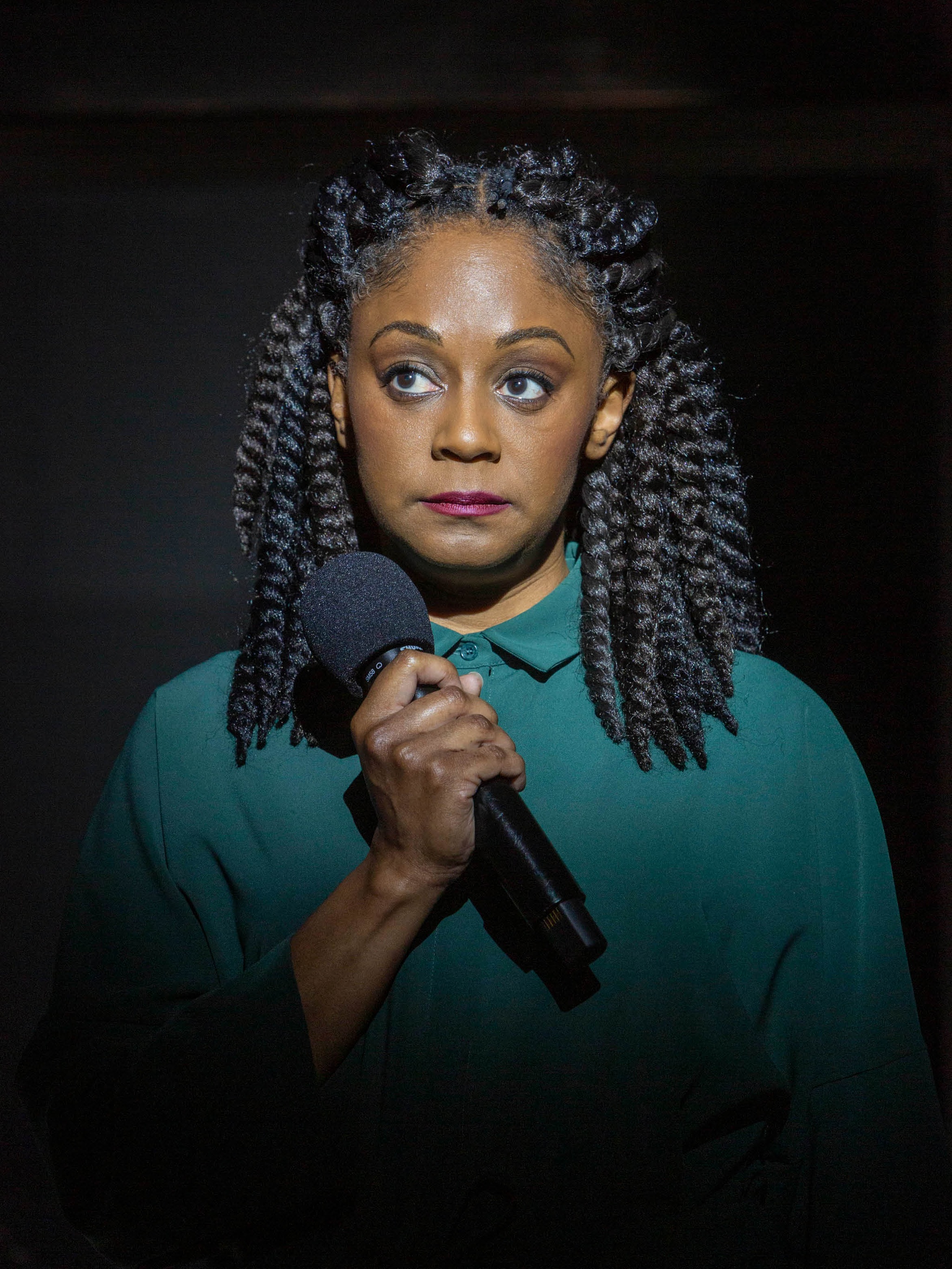 A Black woman wearing a green jumpsuit and standing with her face illuminated by a spotlight grips a microphone
