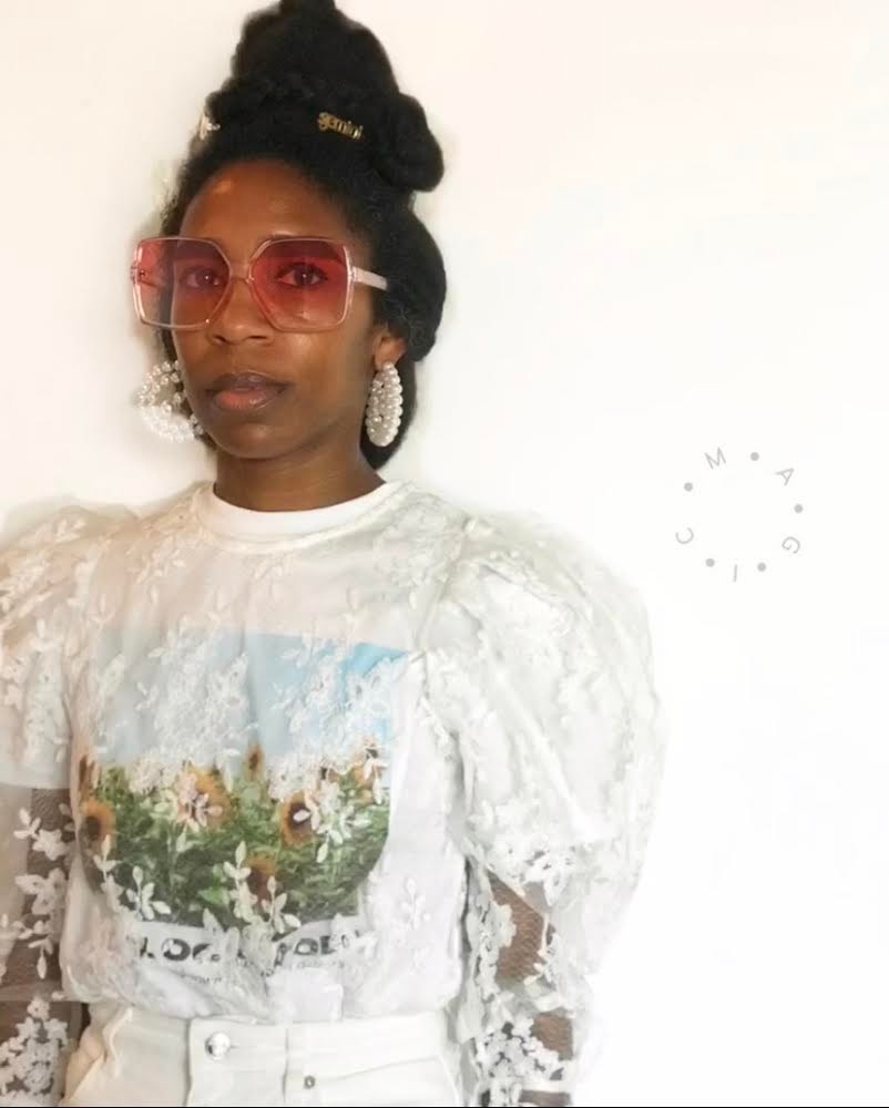 A portrait of DJ April Hunt, a Black woman wearing a white blouse with a garden scene printed on it. She wears rose-tinted glasses and stands against a white wall.