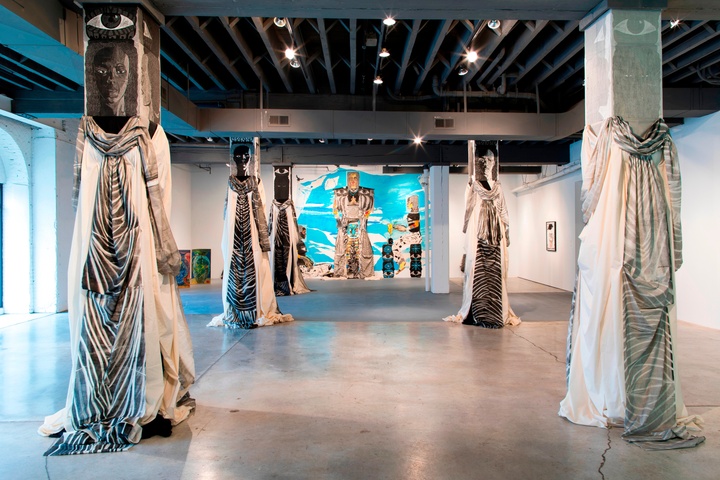 Gallery installation view of works by Paula Wilson, including several pillars draped with fabric, with painted heads to resemble figures, and a painted mural of figures on the back wall.