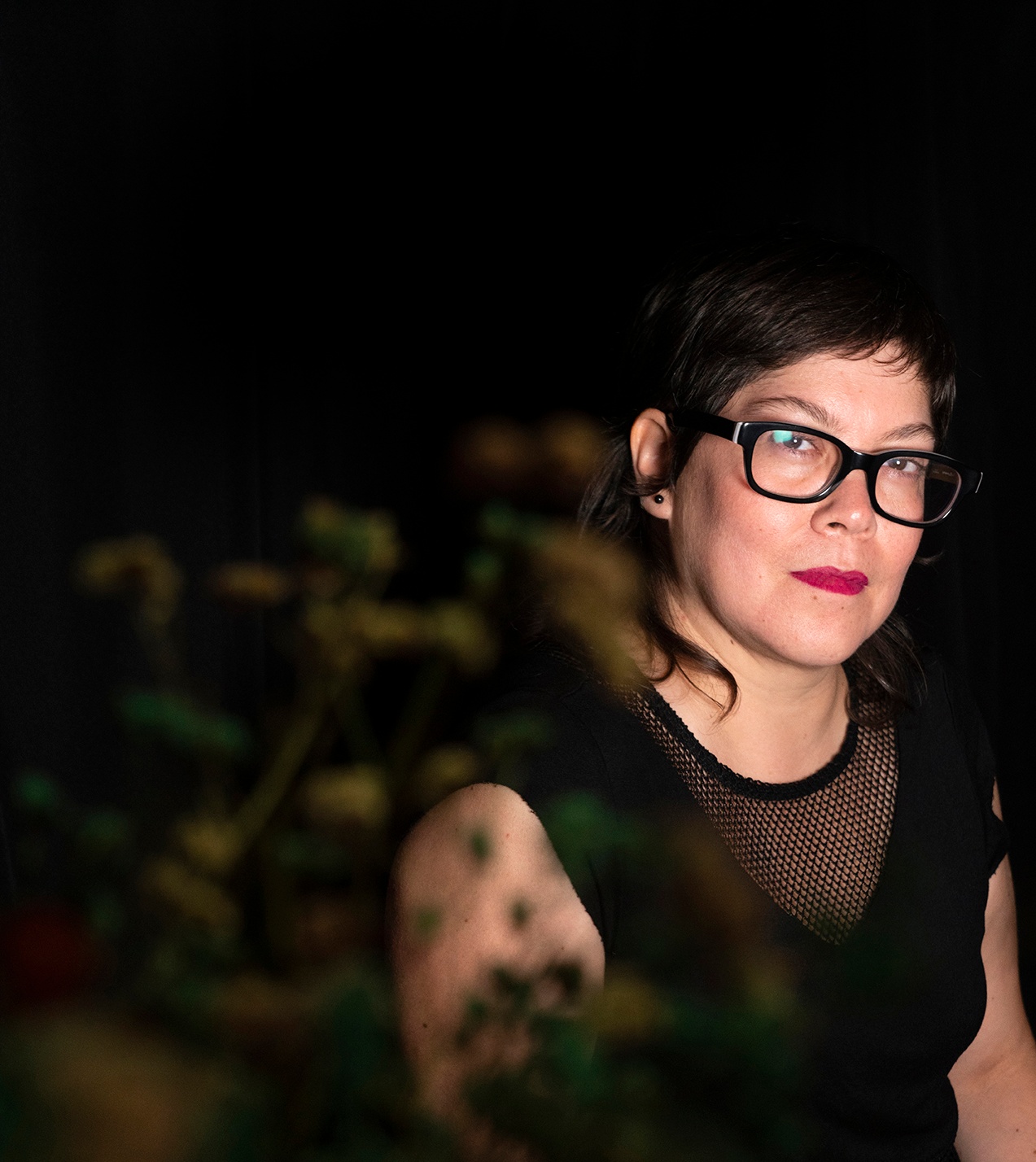 A woman seen through a branch with green leaves that is blurry in the foreground. She has brown hair and wears rectangular glasses, and is well lit by a light shining directly on her with the background in deep shadow.