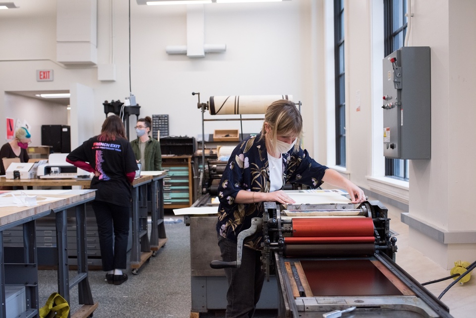 Several people work in a printmaking shop; one operates a roller press. 