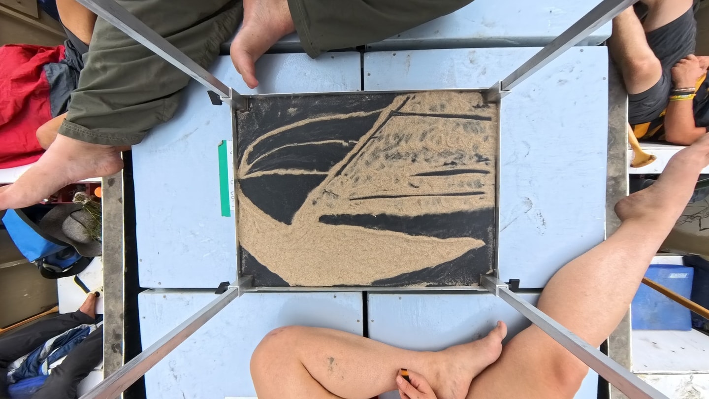 An overhead view of an abstract sand drawing within a silver frame, surrounded by people sitting barefoot on a boat deck.