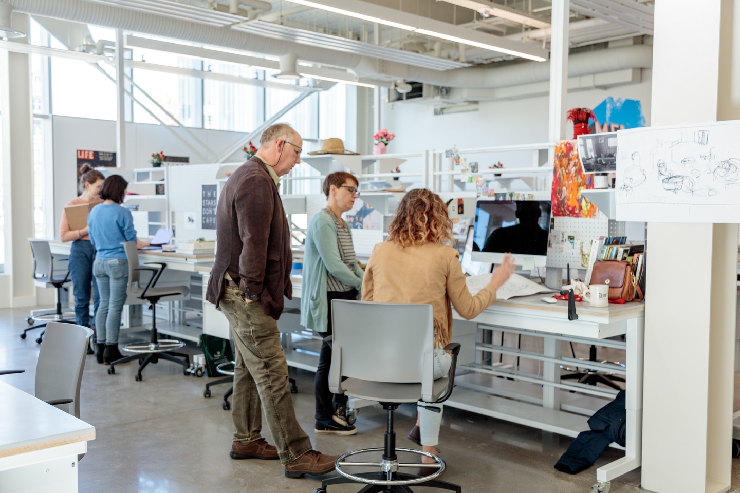 Two faculty gather next to a student at her desk, looking at work; other students are in the background of the light-filled studio space.