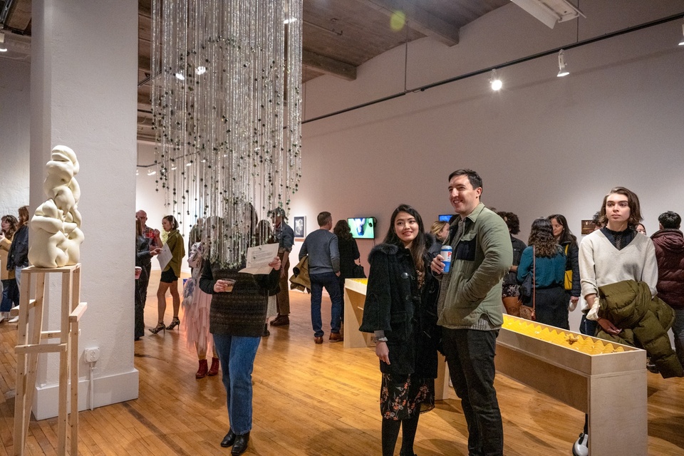 Visitors fill a gallery space. In the foreground is a sculpture of long metallic beaded threads.
