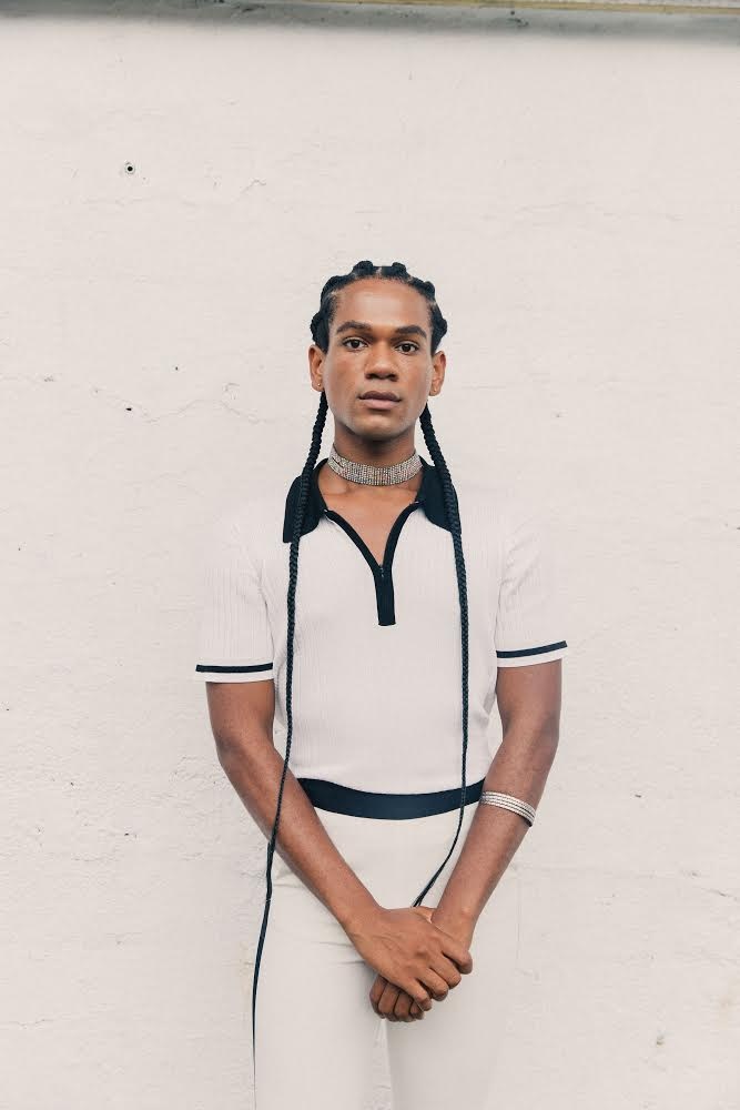 A person with two long braids hanging down below their waist on either side of their body. They stand with arms crossed in front of their body wearing an all-white outfit with a dark collar and bands around the short sleeves and waist of the shirt. 