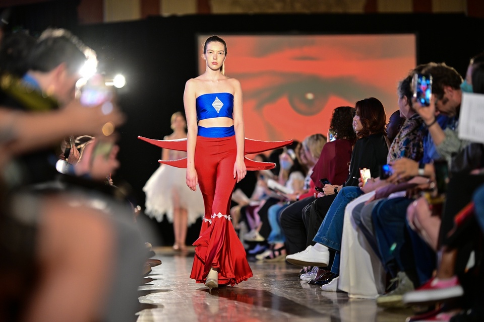 A model walking down the runway in a blue tube top and bell bottom red pants with large wings in red